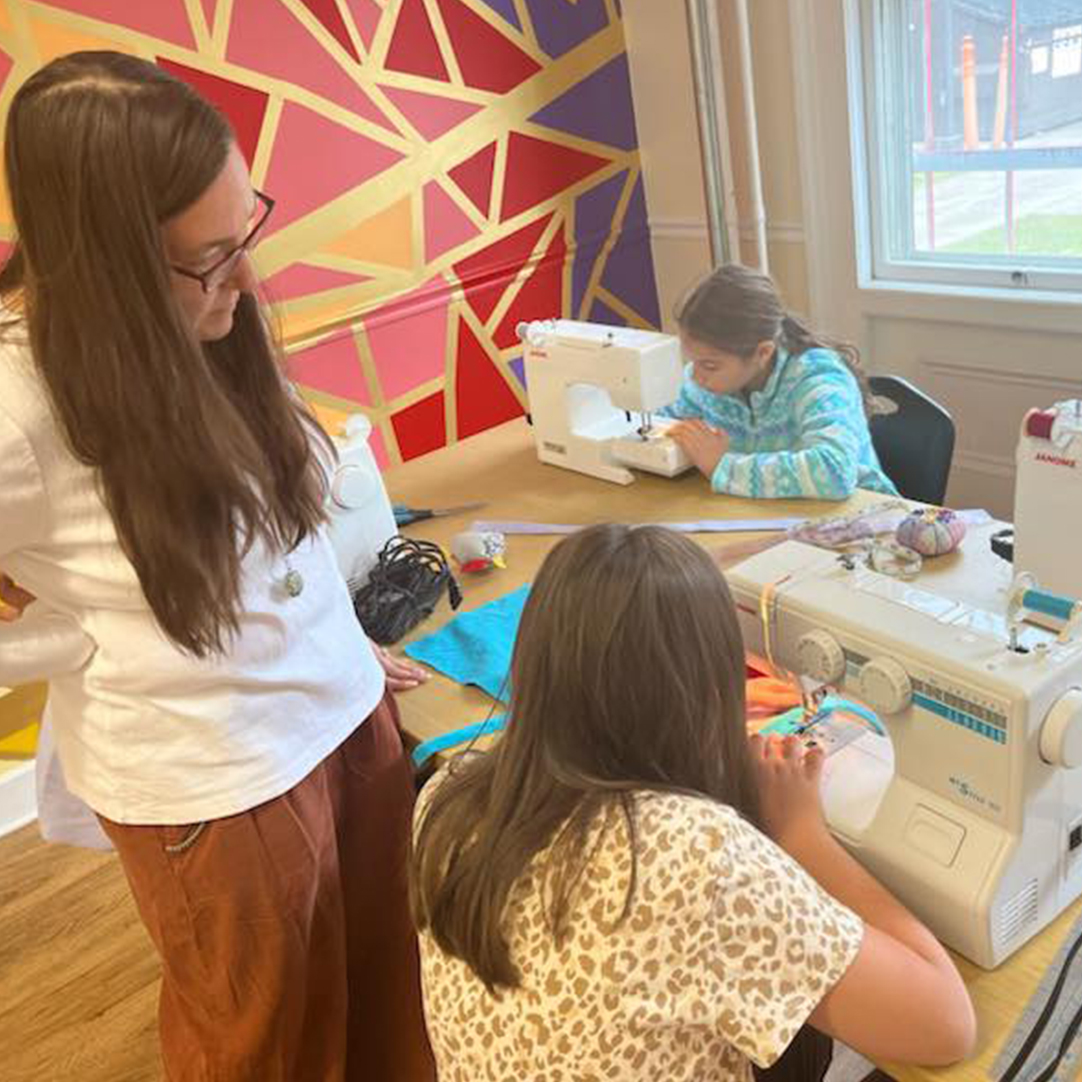 A teacher observing two students working with sewing machines.